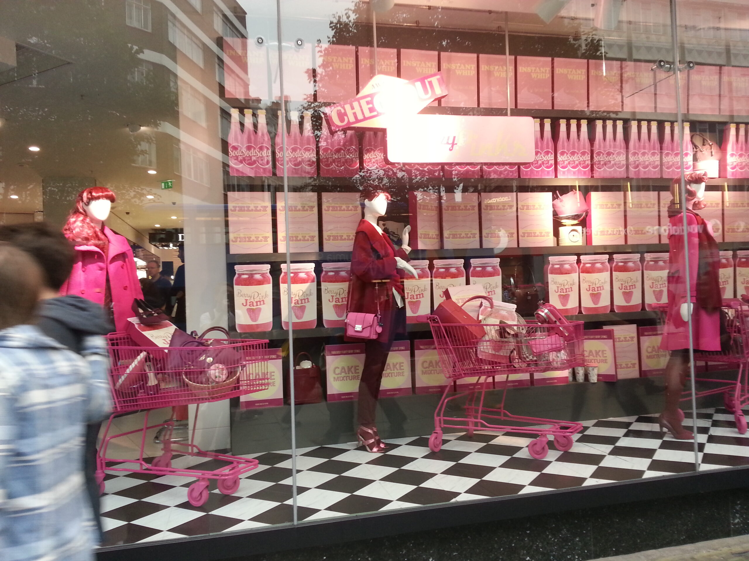 Retail window display with mannequins, pink shopping carts, and oversized food-themed props in pink supermarket setting